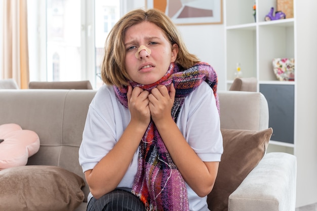 Mujer joven malsana con bufanda caliente alrededor del cuello que se siente mal y enferma que sufre de gripe y resfriado con un parche en la nariz que parece preocupada y confundida sentada en el sofá en la sala de estar iluminada