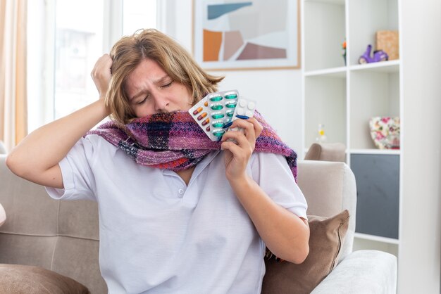 Mujer joven malsana con bufanda caliente alrededor del cuello que se siente mal y enfermo que sufre de gripe y resfriado sosteniendo diferentes pastillas con aspecto preocupado sentado en el sofá en la sala de estar luminosa