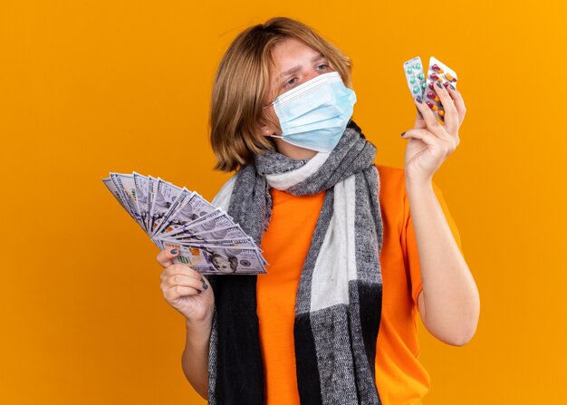 Mujer joven malsana con bufanda caliente alrededor de su cuello con máscara facial protectora sosteniendo pastillas y dinero en efectivo que parece confundido