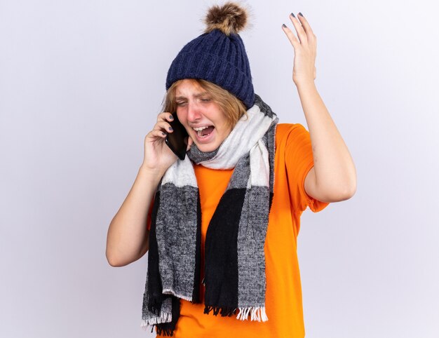 Mujer joven malsana en camiseta naranja con bufanda caliente alrededor del cuello y sombrero sintiéndose terrible sufrimiento de gripe hablando por teléfono móvil con expresión agresiva preocupada en la pared blanca