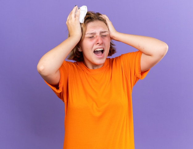 Mujer joven malsana en camiseta naranja que se siente terrible gritando tocando su cabeza que sufre de fiebre y fuerte dolor de cabeza