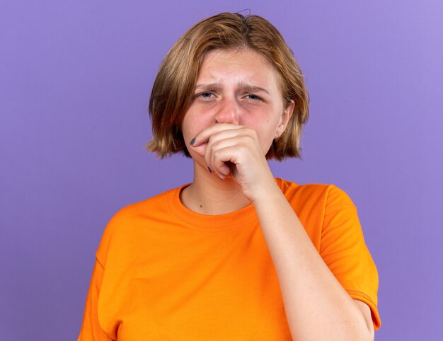 Mujer joven malsana en camiseta naranja que se siente terrible limpiándose la nariz con fiebre resfriado de pie sobre la pared púrpura