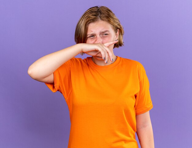 Mujer joven malsana en camiseta naranja que se siente terrible limpiándose la nariz con la mano que sufre de goteo nasal con expresión triste de pie sobre la pared púrpura
