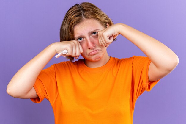 Mujer joven malsana en camiseta naranja que se siente terrible sujetando el tejido que sufre de goteo nasal cought frío llorando duro frotándose los ojos de pie sobre la pared púrpura