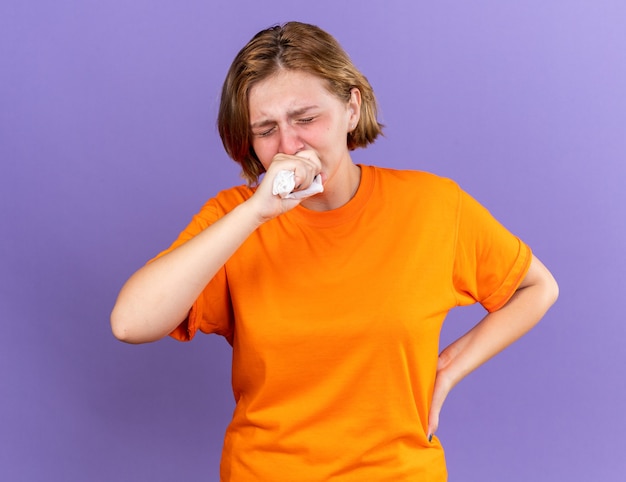 Mujer joven malsana en camiseta naranja que se siente terrible tosiendo en el puño que sufre de virus de pie sobre la pared púrpura