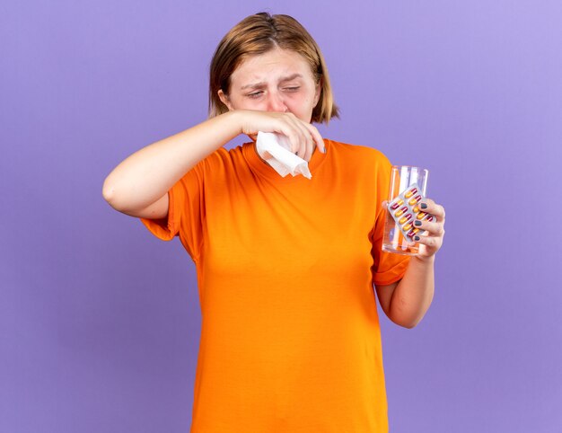 Mujer joven malsana en camiseta naranja con un vaso de agua y pastillas que se siente terrible que sopla la nariz que gotea cogió frío estornudando en un pañuelo de papel de pie sobre una pared púrpura
