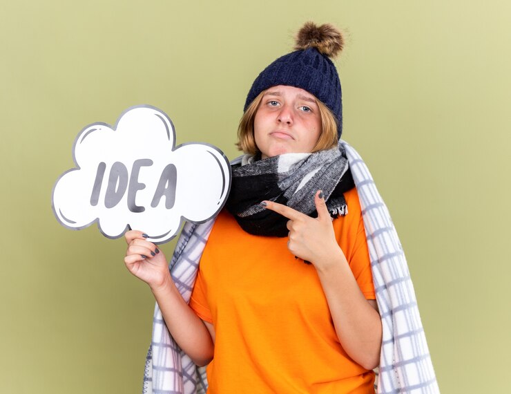 Foto gratuito mujer joven malsana con gorro y bufanda alrededor del cuello envuelta en una manta sintiéndose mal atrapado en frío sosteniendo el letrero de la burbuja del discurso con la idea de la palabra apuntando con el dedo