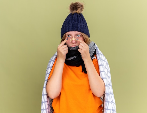 Foto gratuita mujer joven malsana con gorro y bufanda alrededor del cuello envuelta en una manta sintiéndose mal sosteniendo un parche pegado en la nariz con aspecto confundido y preocupado de pie sobre la pared verde