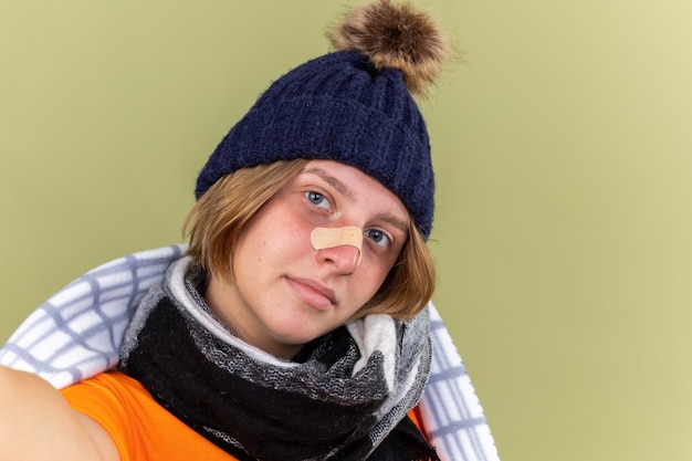 Mujer joven malsana con gorro y bufanda alrededor del cuello envuelto en una manta que sufre de frío con parche en la nariz sonriendo de pie sobre la pared verde