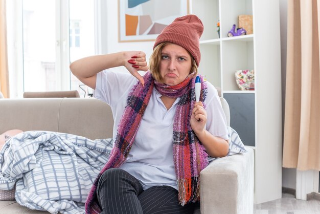 Mujer joven malsana en gorro con bufanda alrededor del cuello sosteniendo el termómetro mostrando los pulgares hacia abajo frunciendo los labios sentado en el sofá en la sala de luz