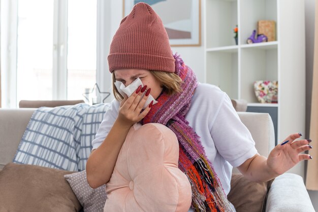 Mujer joven malsana en gorro con bufanda sosteniendo la almohada que sopla la nariz en un tejido que sufre de resfriado y gripe sentado en la silla en la sala de estar ligera