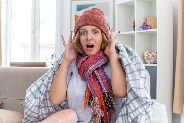 Mujer joven malsana con gorro envuelto en una manta con aspecto de malestar y enfermo que sufre de resfriado y gripe con termómetro con fiebre con aspecto preocupado y asustado sentado en el sofá en la sala de estar luminosa