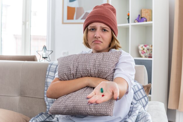 Mujer joven malsana en gorro con manta malestar y malestar que sufre de resfriado y gripe sosteniendo almohada y pastillas preocupado sentado en el sofá en la sala de estar luminosa