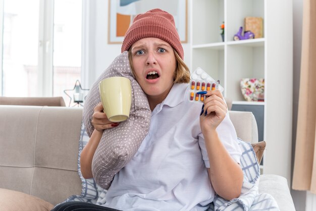 Mujer joven malsana en gorro con manta malestar y malestar que sufren de resfriado y gripe sosteniendo almohada y taza con pastillas preocupado sentado en el sofá en la sala de estar luminosa