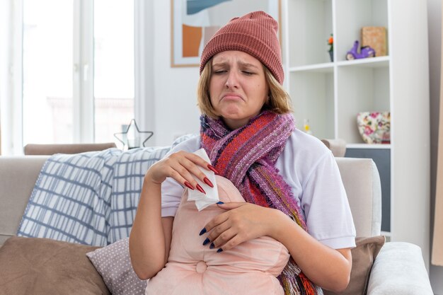 Mujer joven malsana molesta en gorro con bufanda con aspecto de malestar y enfermo sosteniendo tejido que sufre de resfriado y gripe y sonarse la nariz sentado en el sofá en la sala de estar luminosa