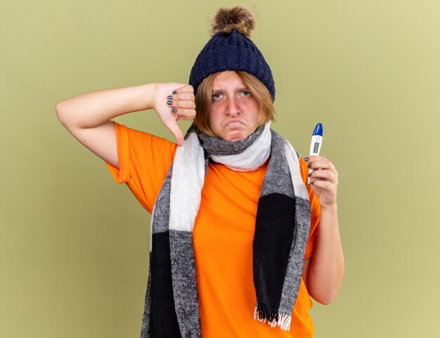 Mujer joven malsana con sombrero con bufanda alrededor de su cuello que se siente mal sosteniendo un termómetro digital que sufre de gripe y fiebre mostrando los pulgares hacia abajo de pie sobre la pared verde