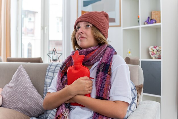 Mujer joven malsana con sombrero con bufanda caliente alrededor del cuello que se siente mal y enfermo que sufre de resfriado y gripe sosteniendo una botella de agua caliente con aspecto preocupado sentado en el sofá en la sala de estar luminosa