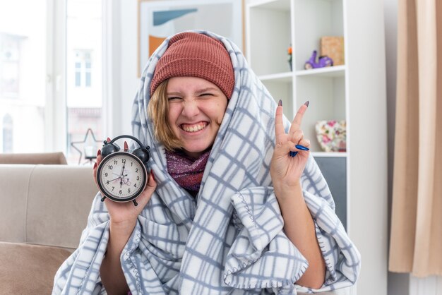 Mujer joven malsana con sombrero envuelto en una manta con reloj despertador sonriendo mostrando signo v sentirse mejor sentado en el sofá en la sala de luz