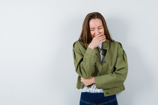 Mujer joven con la mano en la boca en camisa, chaqueta y mirando alegre, vista frontal.