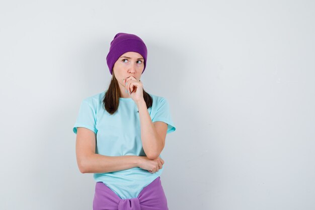 Mujer joven con la mano en la boca, pensando en algo en camiseta azul, gorro morado y mirando pensativo. vista frontal.