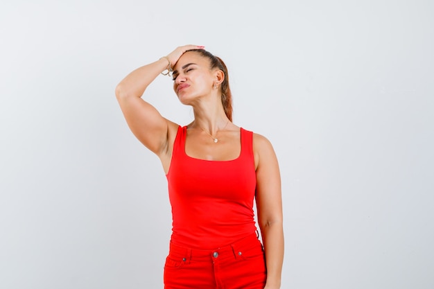 Mujer joven con la mano en la cabeza en camiseta roja, pantalones y mirando doloroso, vista frontal