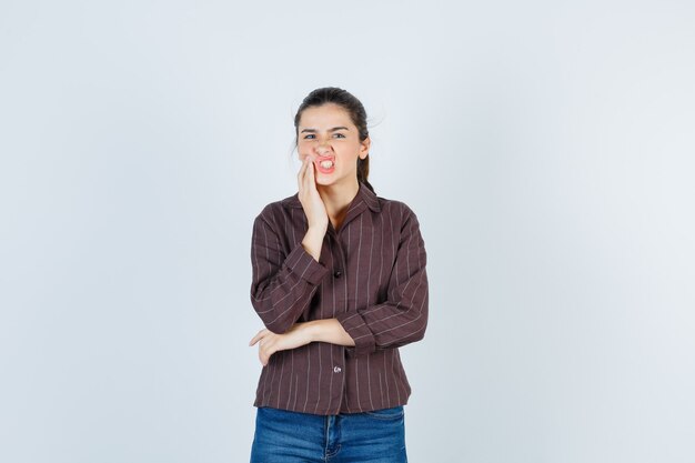 Mujer joven con la mano cerca de la boca, con dolor de muelas en camisa a rayas, jeans y luciendo exhausta. vista frontal.