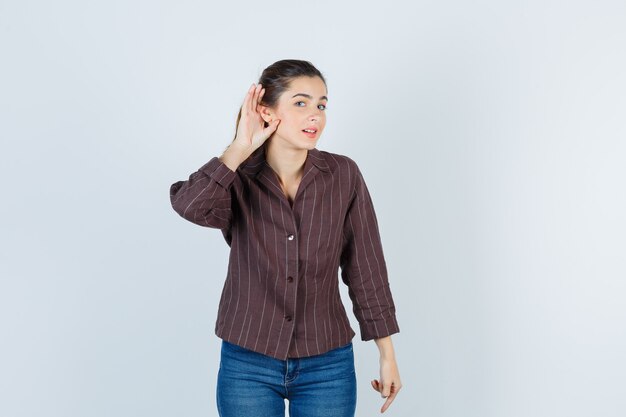 Mujer joven con la mano cerca de la oreja para escuchar algo en camisa a rayas, jeans y mirando enfocado, vista frontal.