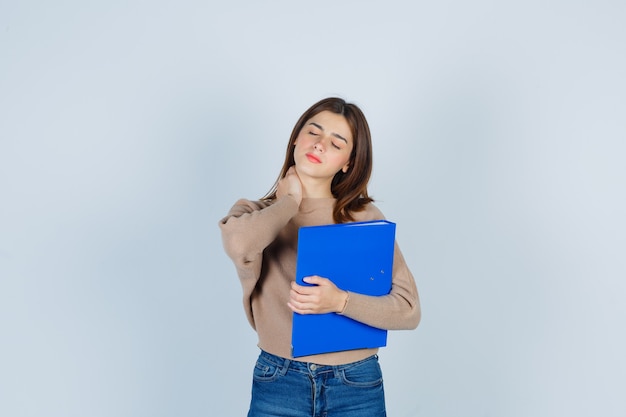 Mujer joven con la mano en el cuello, manteniendo la carpeta en suéter, jeans y luciendo dolorosa, vista frontal.