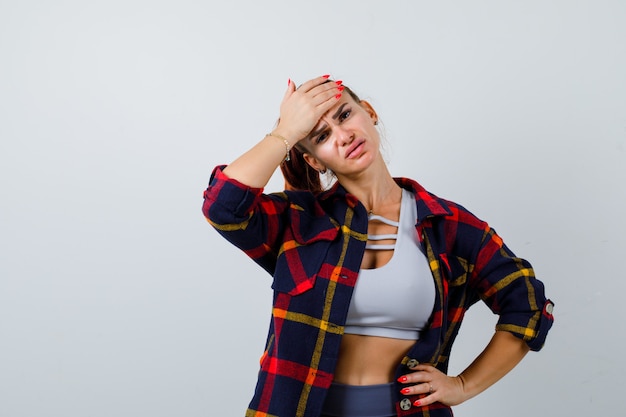 Mujer joven con la mano en la frente mientras mantiene la mano en la cadera en la parte superior de la cosecha, camisa a cuadros, pantalones y con aspecto cansado, vista frontal.