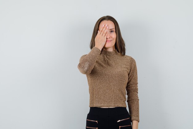 Mujer joven con la mano en el ojo en blusa dorada, vista frontal.