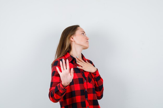Mujer joven con la mano sobre el pecho, mostrando la señal de pare en camisa a cuadros y luciendo agitada. vista frontal.