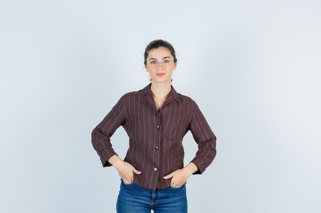 Mujer joven con las manos en los bolsillos en camisa a rayas, jeans y mirando feliz, vista frontal.