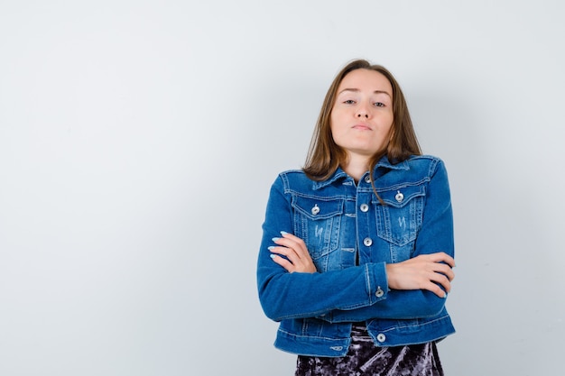 Foto gratuita mujer joven con las manos cruzadas en chaqueta de mezclilla, vestido y mirando confiado, vista frontal.