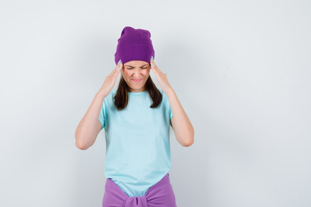 Mujer joven con las manos en las sienes, haciendo muecas en camiseta azul, gorro morado y mirando acosado, vista frontal.