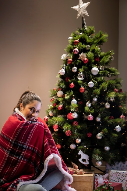 Mujer joven con una manta sentada al lado del árbol de navidad