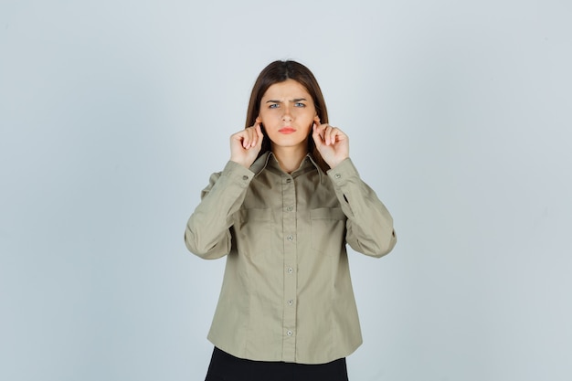 Mujer joven manteniendo los dedos detrás de las orejas en camisa, falda y luciendo sombrío. vista frontal.