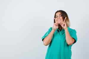 Foto gratuita mujer joven manteniendo los dedos en las mejillas en camiseta de polo y mirando alegre, vista frontal.