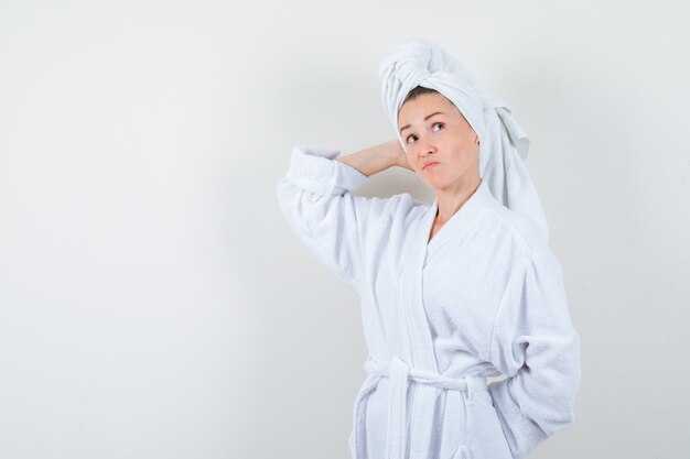 Mujer joven manteniendo la mano en la cabeza en bata de baño blanca, toalla y mirando indeciso, vista frontal.