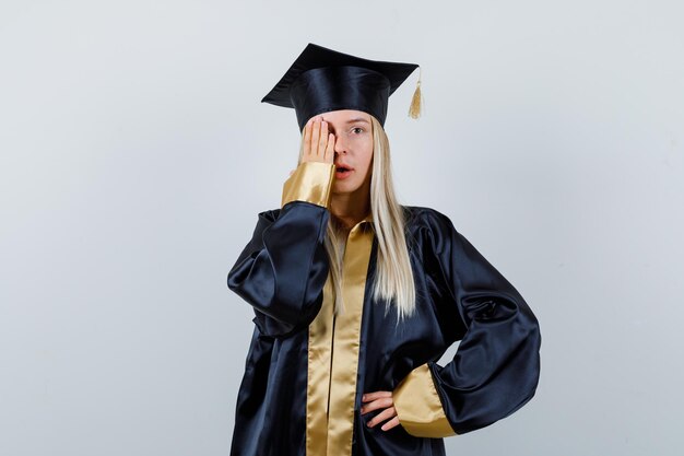 Mujer joven manteniendo la mano en el ojo en uniforme de posgrado y mirando enfocado