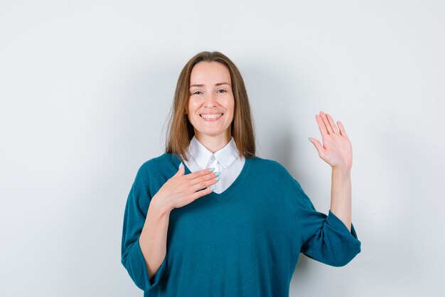 Mujer joven manteniendo la mano en el pecho, mostrando la palma en suéter sobre camisa blanca y mirando alegre. vista frontal.