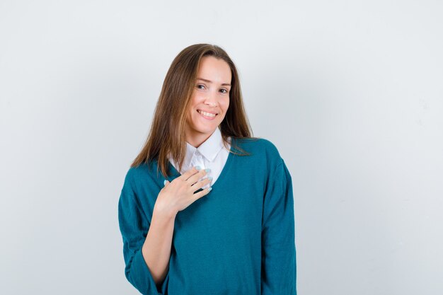 Mujer joven manteniendo la mano en el pecho en suéter sobre camisa blanca y luciendo alegre. vista frontal.