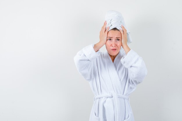 Mujer joven manteniendo las manos en la cabeza en bata de baño blanca, toalla y mirando melancólica. vista frontal.