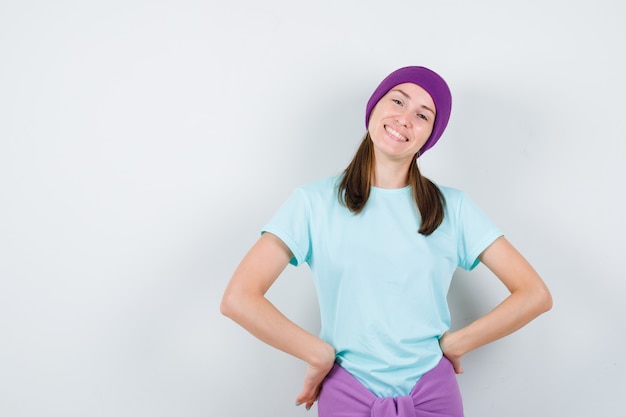 Mujer joven manteniendo las manos en las caderas en camiseta, gorro y mirando feliz. vista frontal.