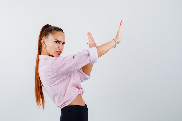 Mujer joven manteniendo las manos para defenderse en camisa casual, pantalones y mirando seria