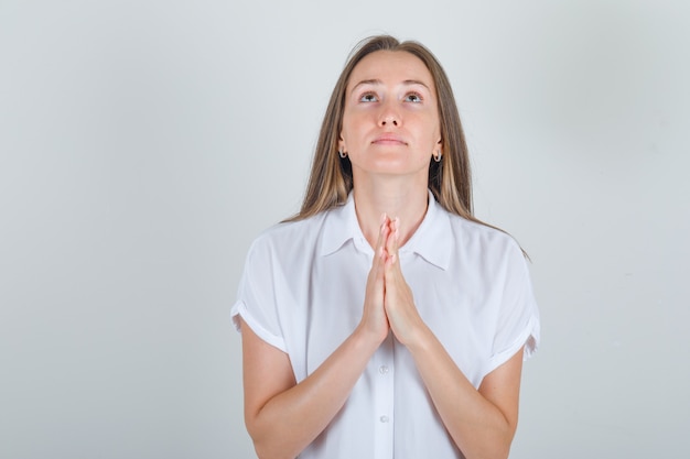 Mujer joven manteniendo las manos en gesto de oración en camisa blanca y mirando esperanzado