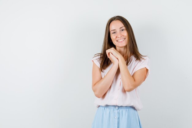 Mujer joven manteniendo las manos juntas en camiseta, falda y luciendo linda vista frontal.