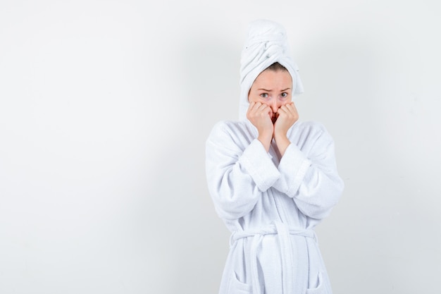 Mujer joven manteniendo los puños en las mejillas en bata de baño blanca, toalla y mirando asustado, vista frontal.