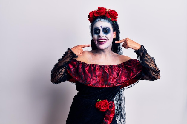 Mujer joven con maquillaje mexicano del día de los muertos sonriendo alegre mostrando y señalando con los dedos los dientes y la boca concepto de salud dental