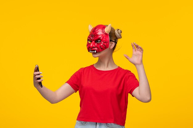 Mujer joven con máscara de diablo aterrador usando su teléfono fondo amarillo halloween