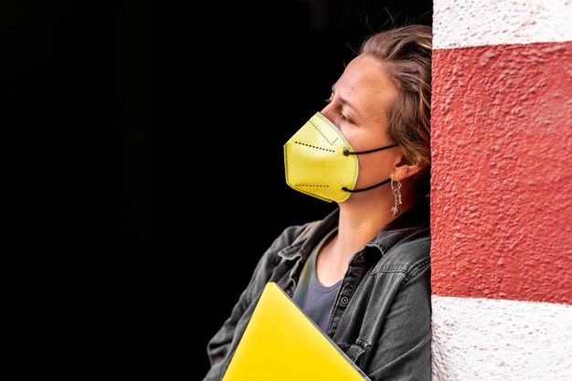 Mujer joven con una máscara facial amarilla y sosteniendo una computadora portátil apoyada en una pared